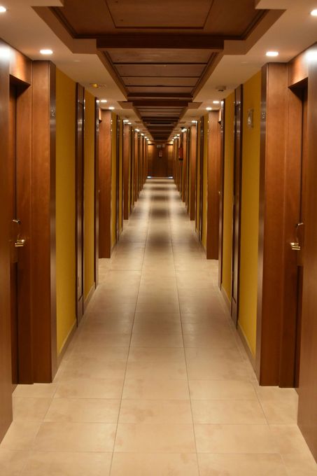 A long, narrow hotel hallway with tiled floors, yellow walls, and wooden doors lining both sides under recessed lighting.