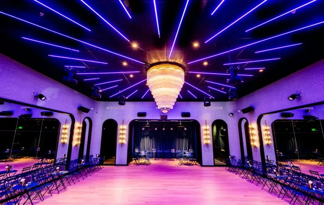 Elegant event space with purple and blue lighting, chandelier, and a stage. Empty folding chairs line the room.