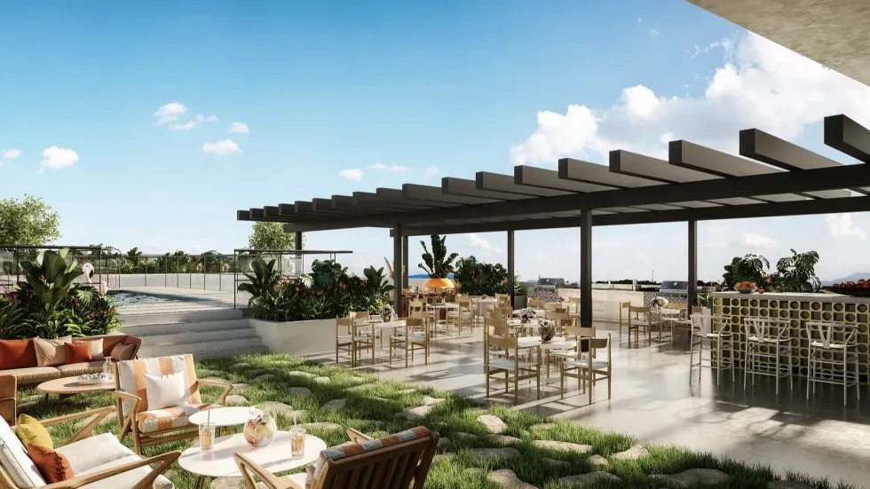 Outdoor patio with tables, chairs, and bar under a pergola; lush greenery, blue sky.