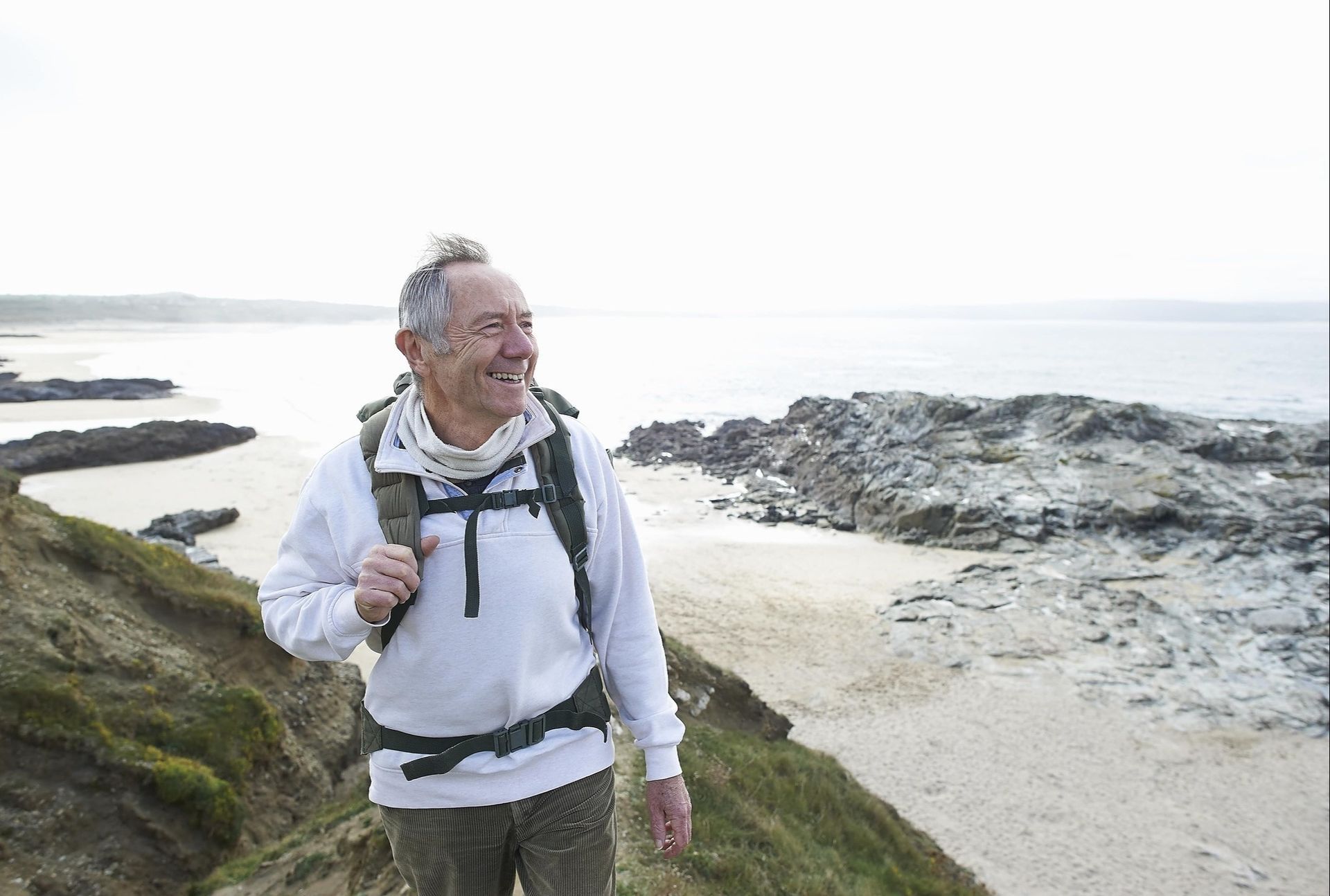 An older man with a backpack is walking on a beach.