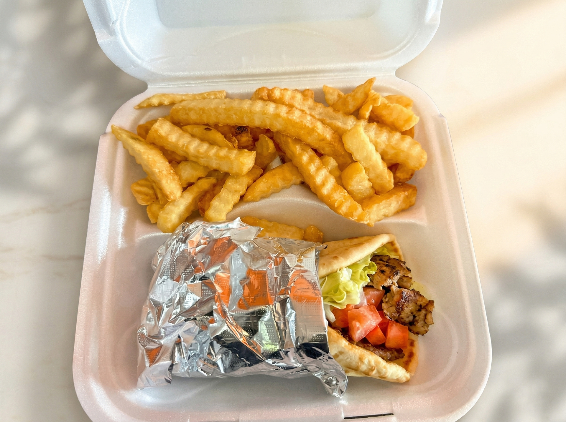 A Styrofoam takeout container with crinkle-cut fries and a foil-wrapped gyro with lettuce, tomatoes, and meat.