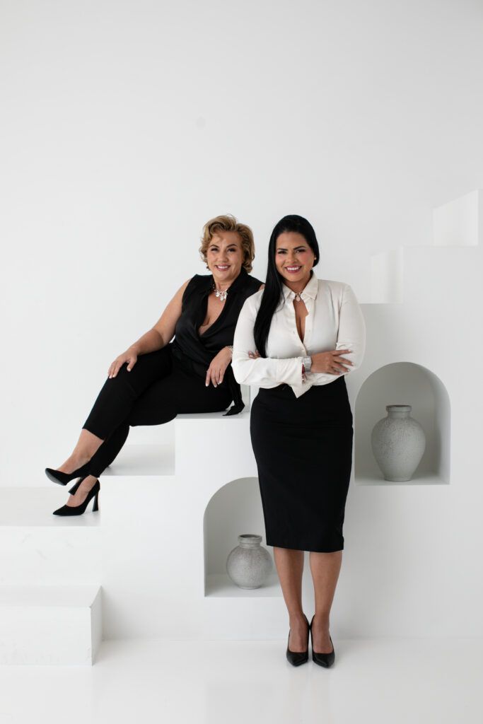 Two women in black uniforms seated, reading magazines, smiling, indoors.