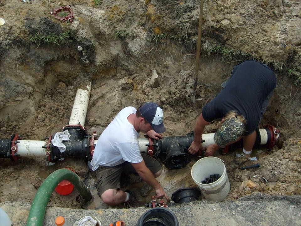 Two plumbers working on pipes in an excavated ditch; one white shirt, one black shirt.