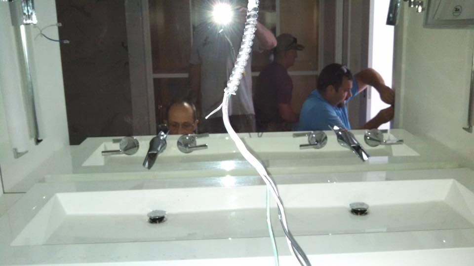 Bathroom with double sinks; people working in the background. Bright lighting, white surfaces, and silver fixtures.