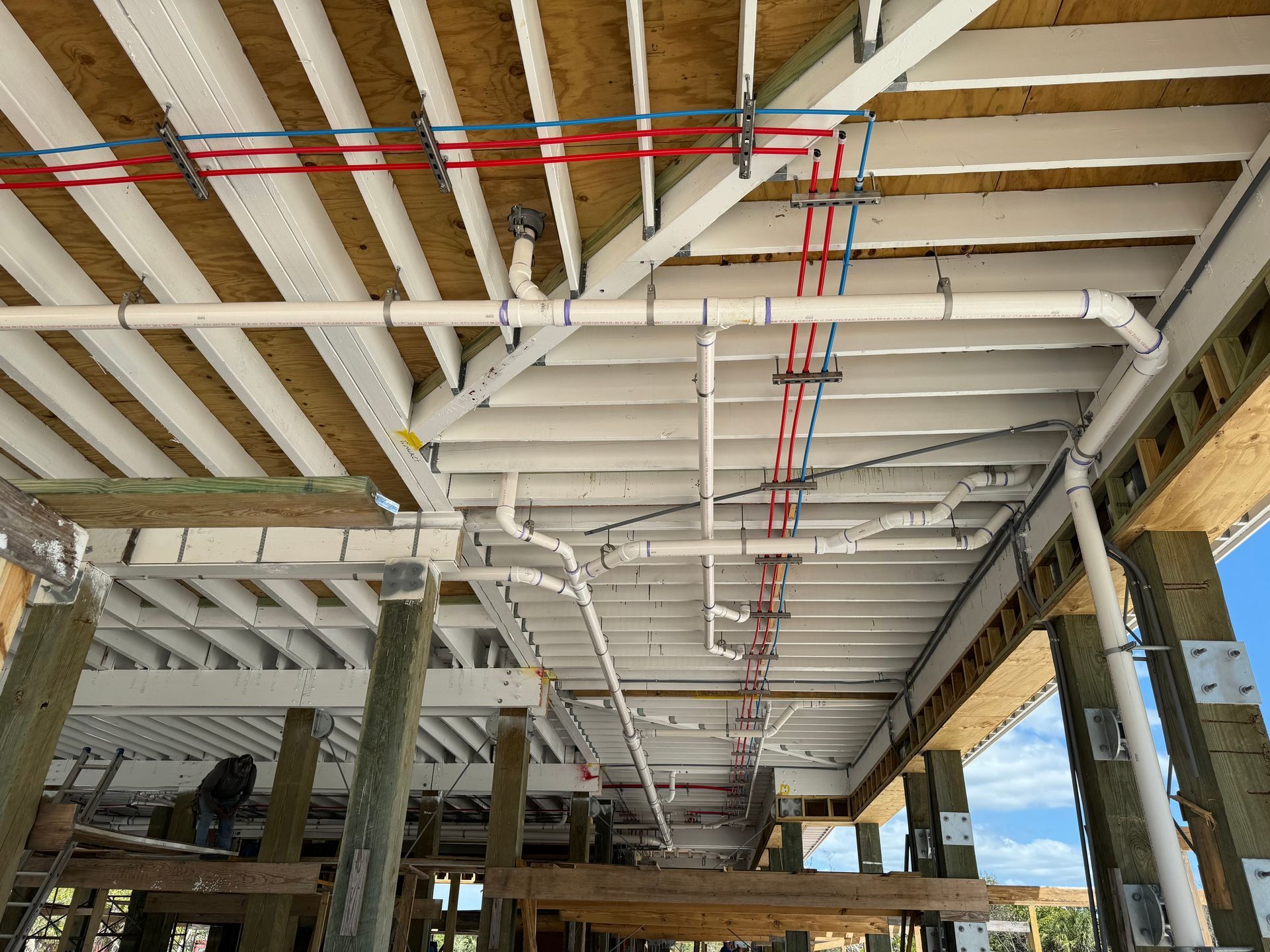 Exposed ceiling beams and pipes in a building under construction, with red and blue conduit overhead.
