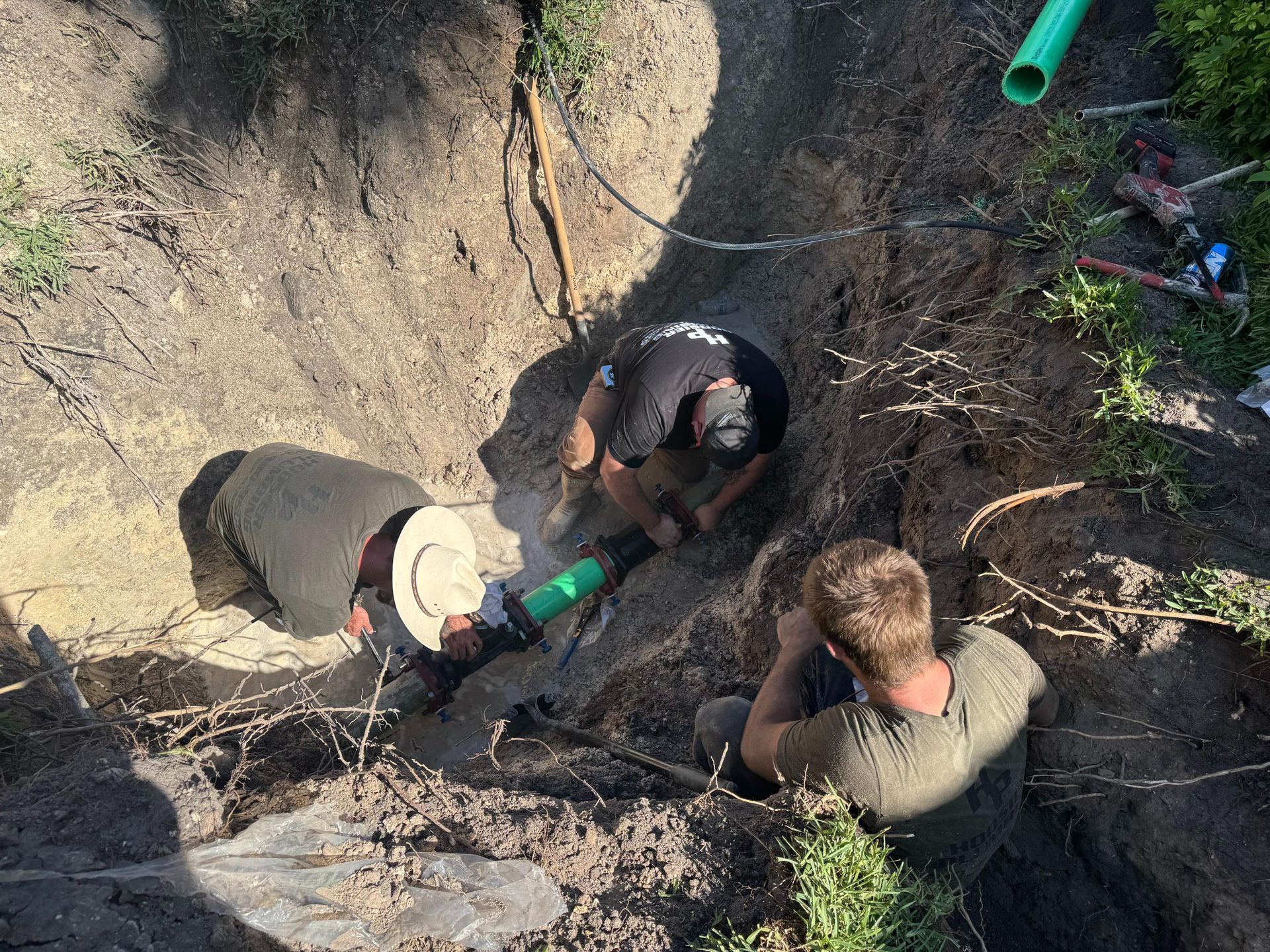 Three workers crouch in a dirt trench, handling a green pipe during excavation.