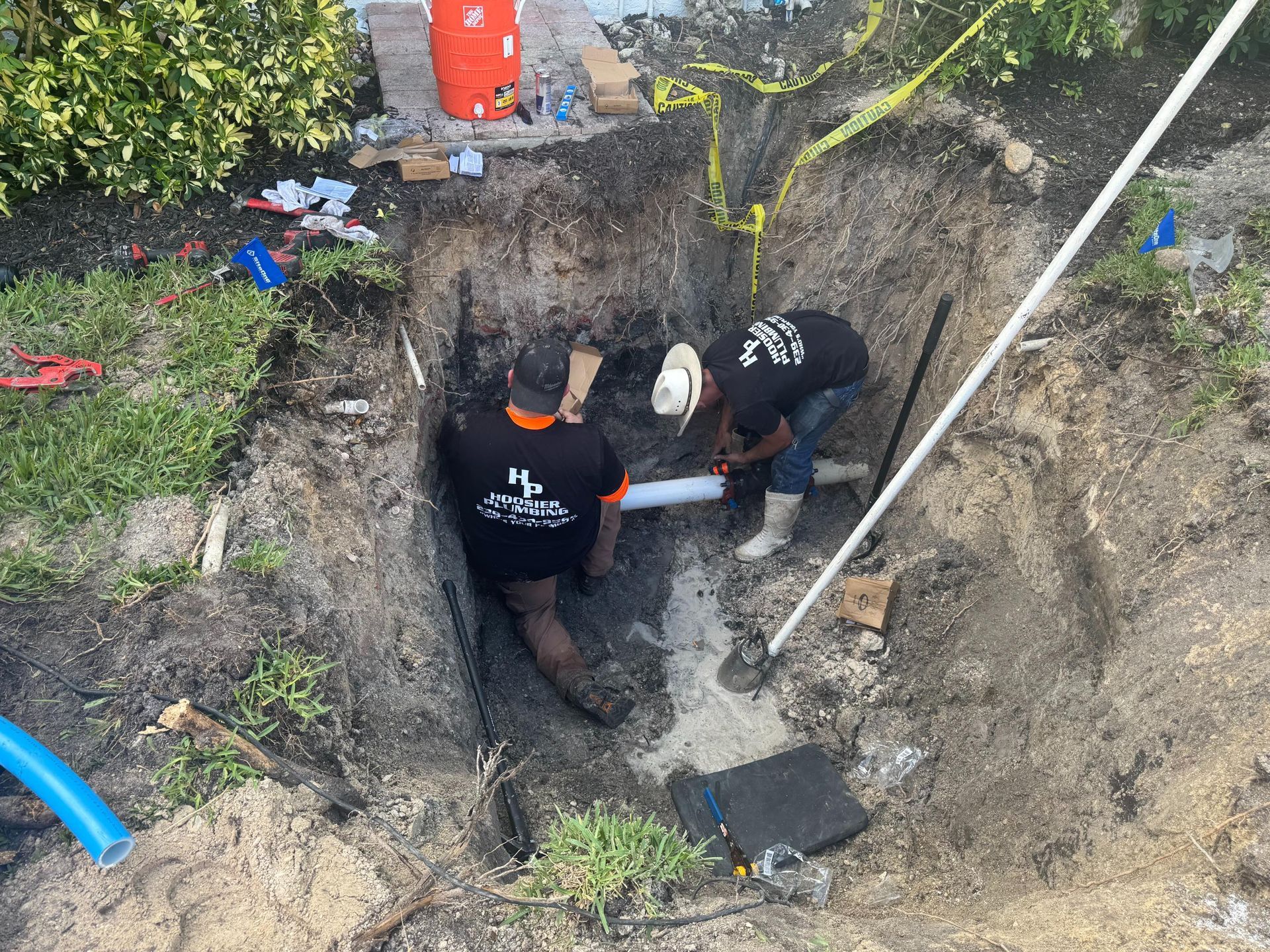 Two workers in a dirt trench repairing underground pipes beside a building wall