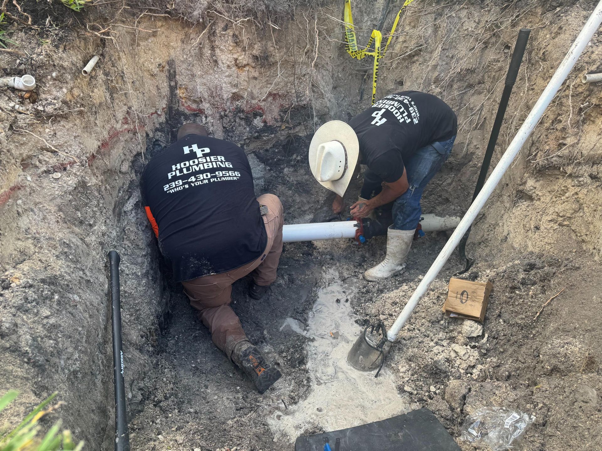 Two workers crouch in a dirt trench, fitting a white pipe beside exposed utility lines.