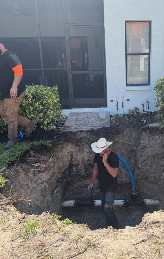 Worker in a white hat sits in a large backyard excavation beside a house with a black sliding door.