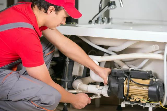 Plumber in red shirt and gray overalls working on pipes under a sink in a bathroom.