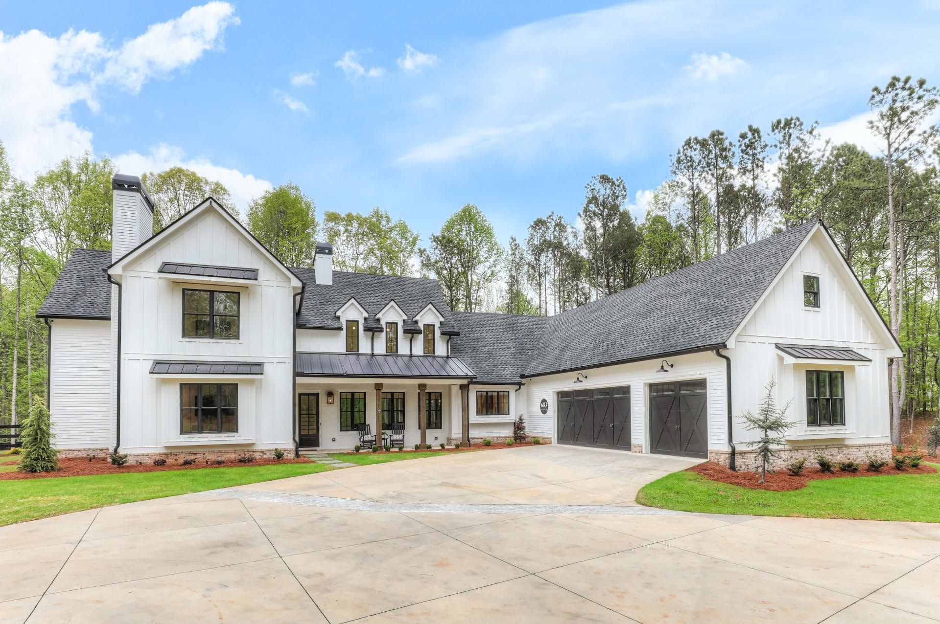 Large white modern farmhouse with stone accents and a wide driveway, set among trees under a blue sky