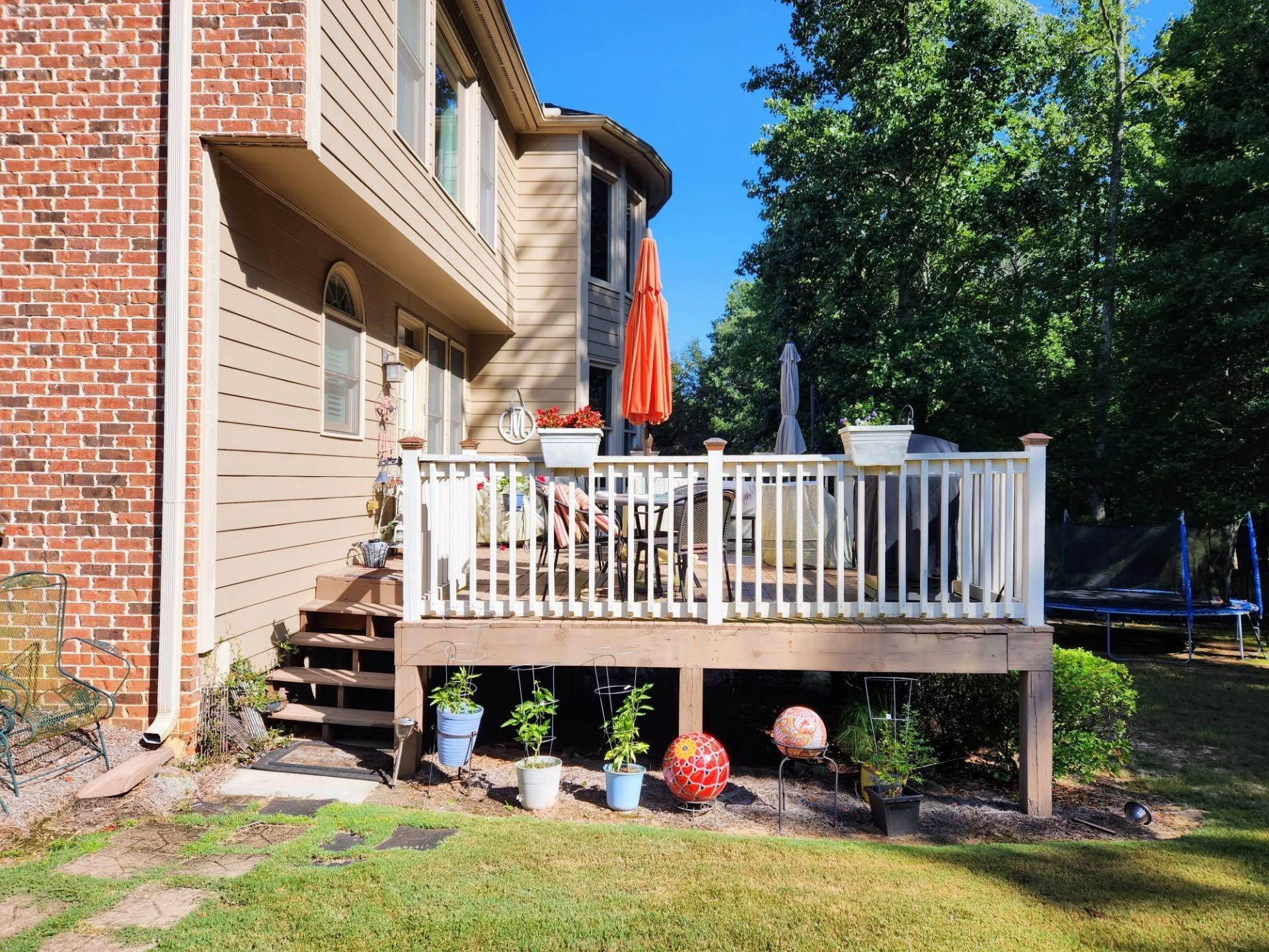 Backyard deck beside brick house with white railing, potted plants, and orange umbrella in sunlight