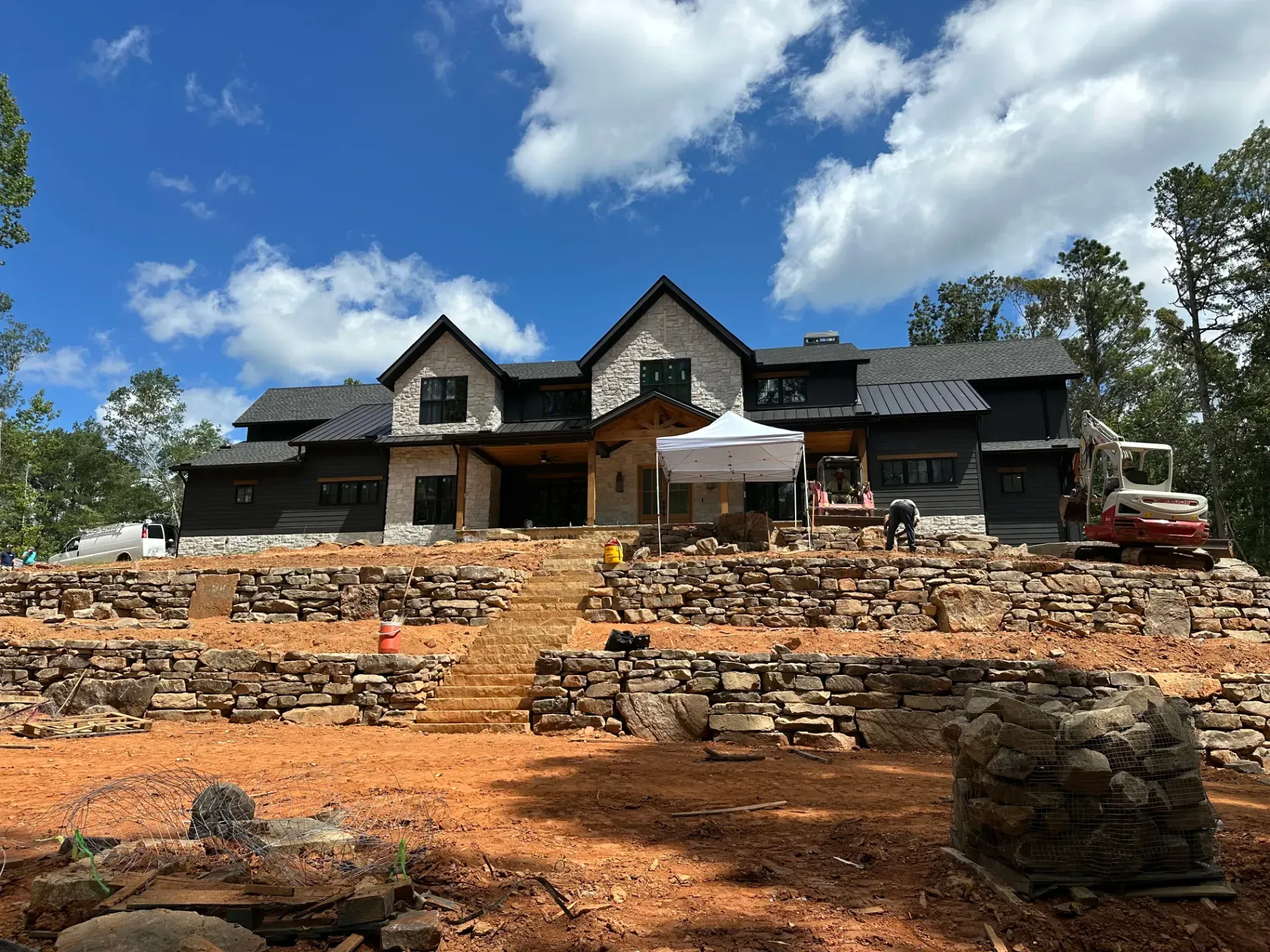 Large stone house under construction on a terraced hillside with cloudy blue sky above