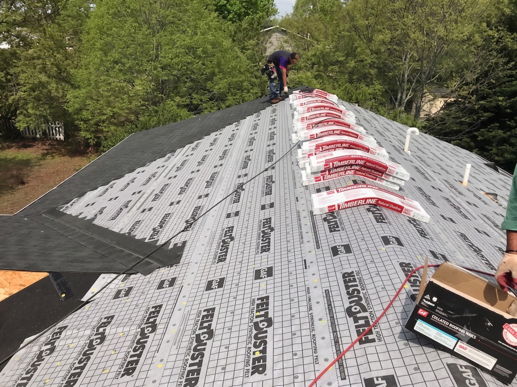 Worker installing roofing underlayment on a steep roof, with trees in the background.