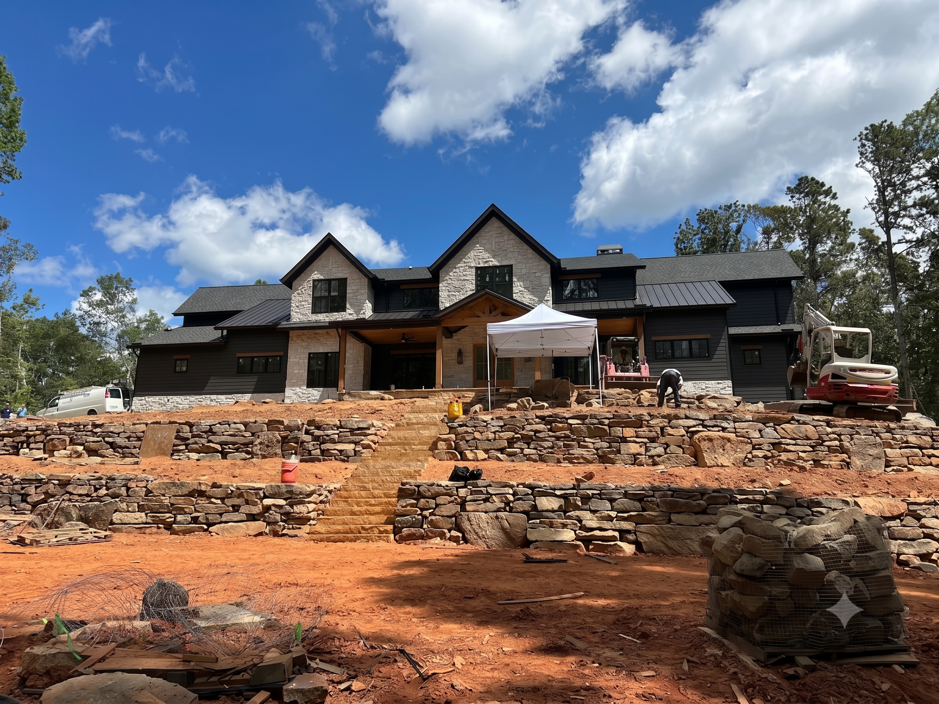 Large dark-stone house under construction on a red dirt lot, with stone retaining walls and a partly cloudy sky