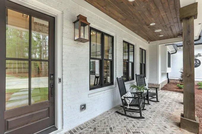Front porch with dark door, black-framed windows, wall lanterns, and two rocking chairs on a brick floor