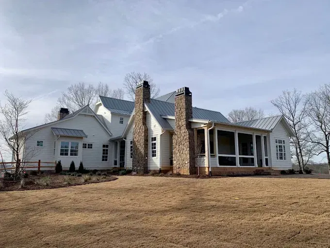 Large white house with stone chimneys and a covered porch on a bare lawn under a cloudy sky