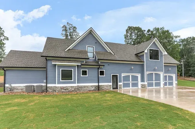 Blue-gray house with white trim, multiple garages, and a large green lawn under a partly cloudy sky