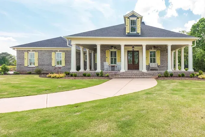 Single-story brick house with white columns, yellow shutters, and a curved driveway on a green lawn