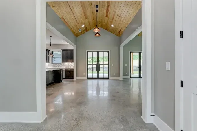 Modern open-plan living room with vaulted wood ceiling, concrete floor, and glass double doors to outside.