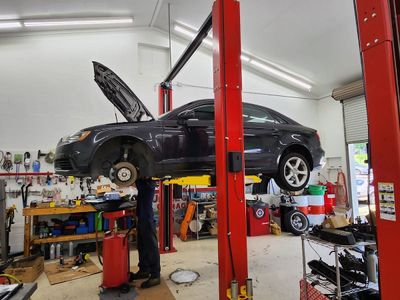 A mechanic works under a dark sedan elevated on a red two-post lift in a well-lit auto repair shop. | Mountain European Auto Repair