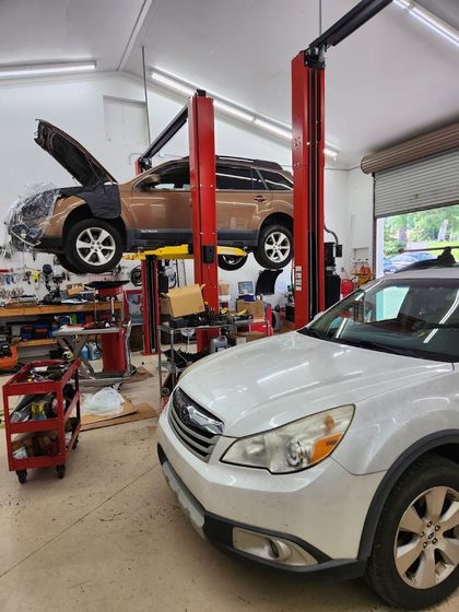 A brown Subaru on a mechanic's lift with its hood open in a garage, next to a white Subaru parked on the floor. | Mountain European Auto Repair