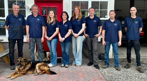 Five people stand in a line posing for a photo in front of a building with a bright red door and windows. | Mountain European Auto Repair