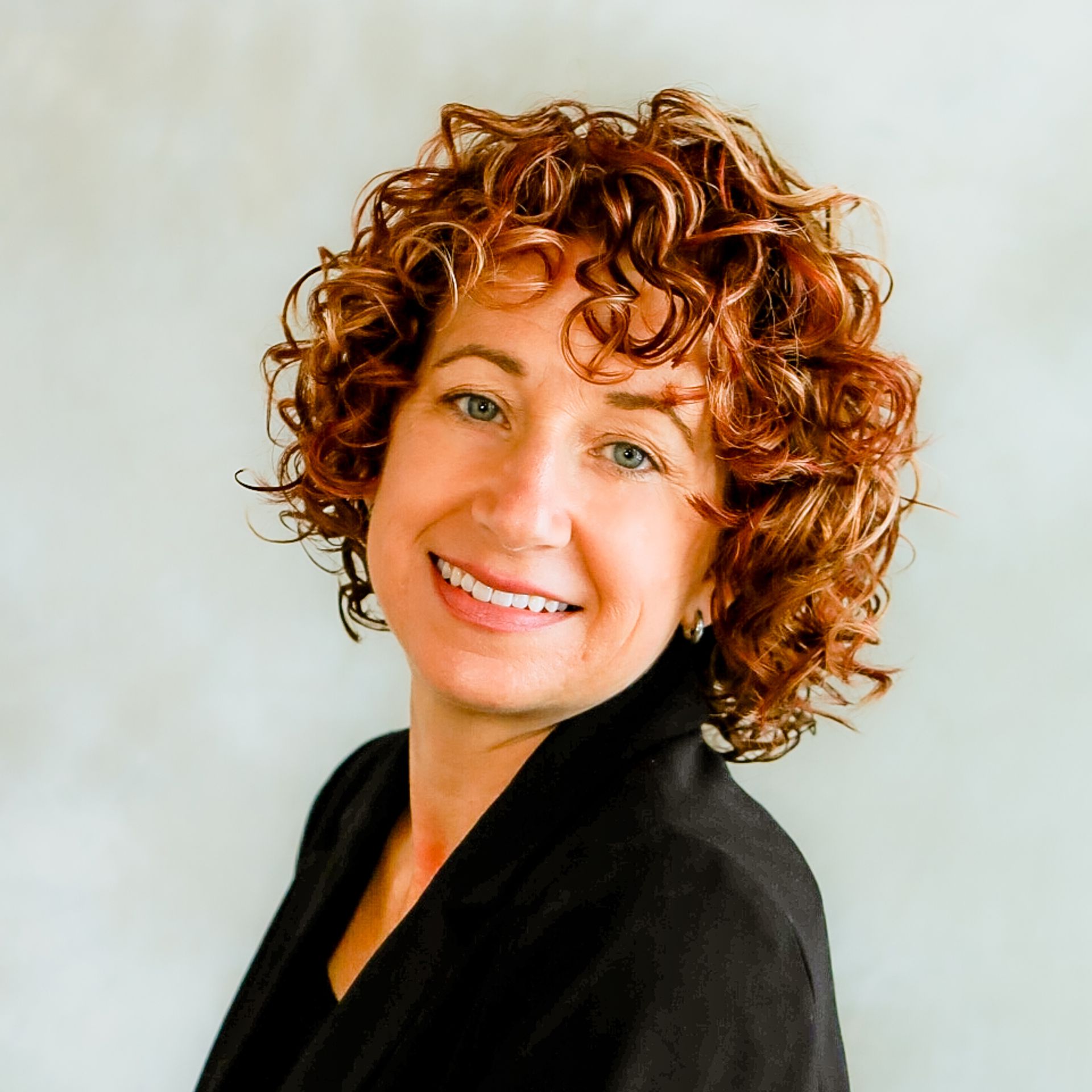 Smiling person with curly auburn hair in a black top against a light gray background