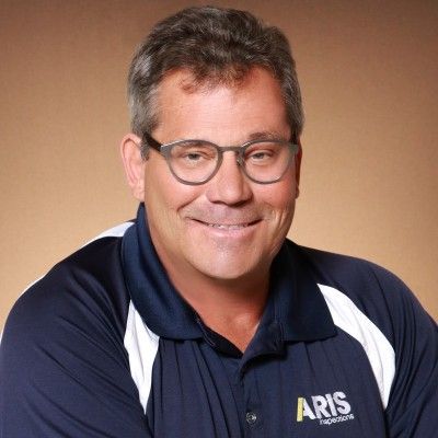 Portrait of a smiling man in glasses wearing a navy polo with ARIS logo on a beige background