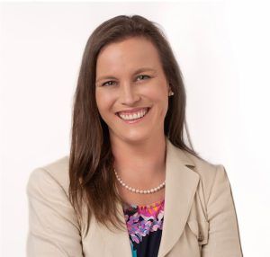A smiling professional wearing a tan blazer over a multi-colored floral top and a pearl necklace against a white background.