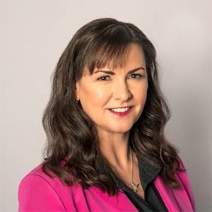 A smiling professional wearing a tan blazer over a multi-colored floral top and a pearl necklace against a white background.