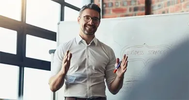 A person in a white button-down shirt stands before a whiteboard, gesturing while presenting in an office setting.