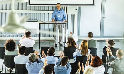 A presenter stands at a lectern before an audience in a bright conference room, with several people raising their hands.