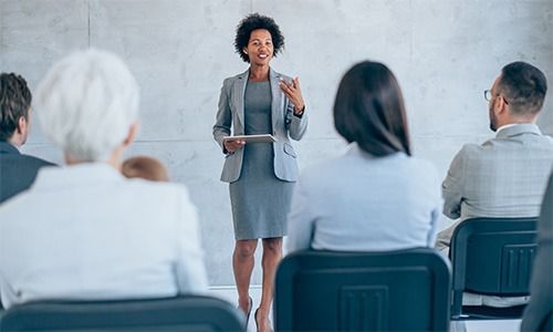 A professional presenting to an audience seated in a bright, modern meeting room.