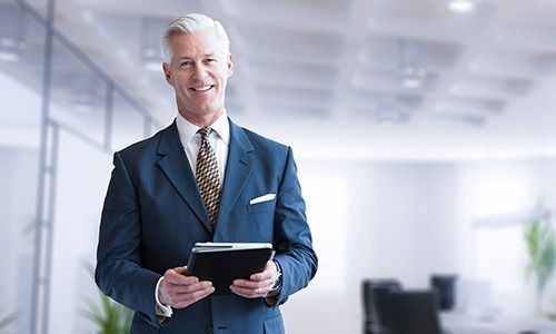 A professional in a blue suit and patterned tie smiling while holding a black tablet in a bright office.