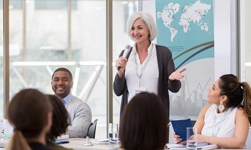 A professional speaker addresses a small meeting group in a bright, modern office with a global map backdrop.
