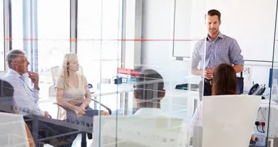 A man standing and presenting to a group of colleagues sitting around a table in a modern, glass-walled office.