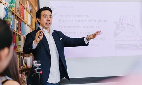 A person in a dark blazer and white shirt gestures while speaking to an audience in front of a presentation screen.