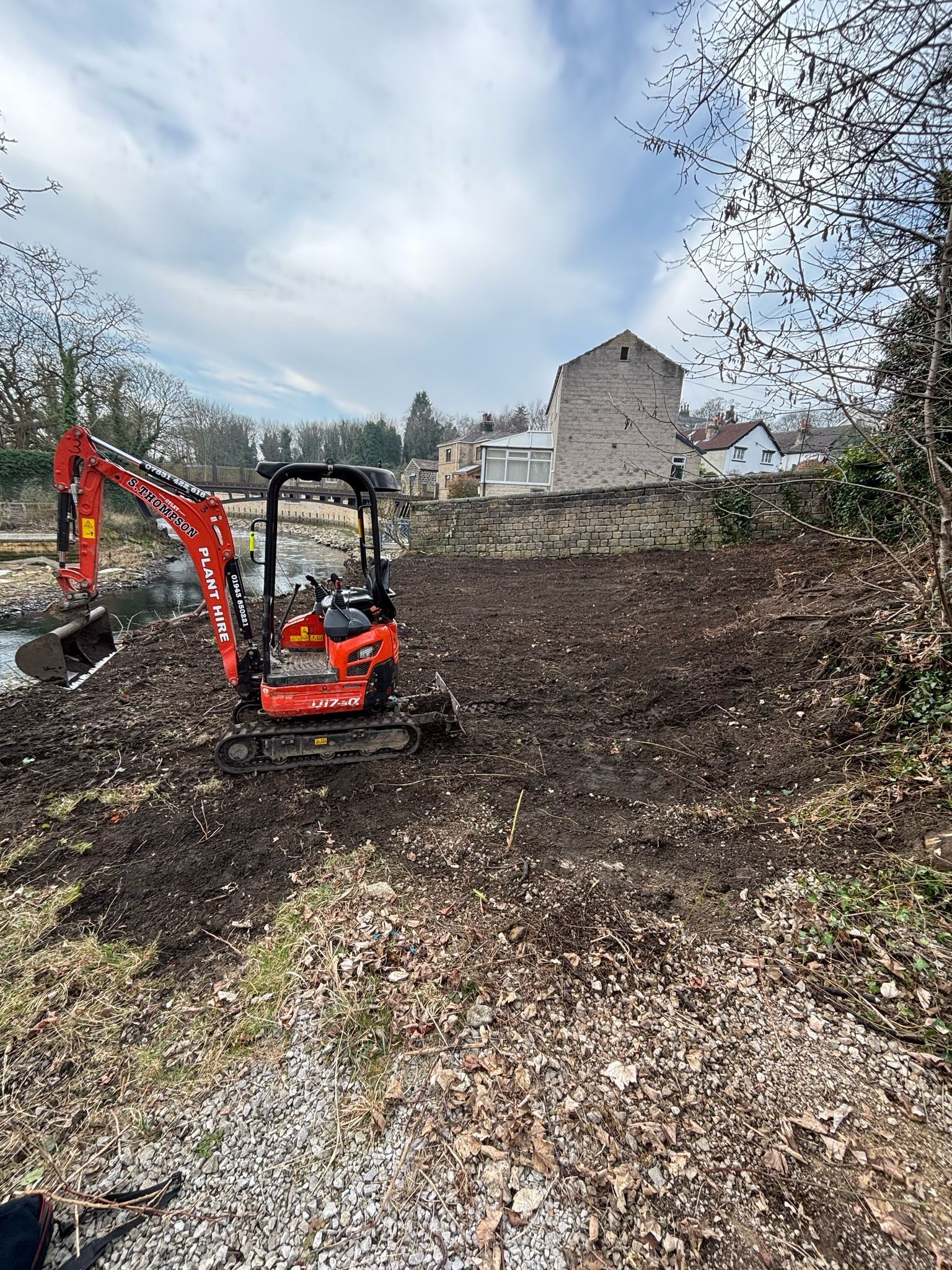 Orange excavator clearing dirt in a muddy yard near a house under a cloudy sky