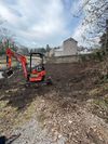 Orange excavator clearing dirt in a muddy yard near a house under a cloudy sky