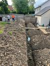 Backyard trench with piles of dirt, workers, and a house beside a fence