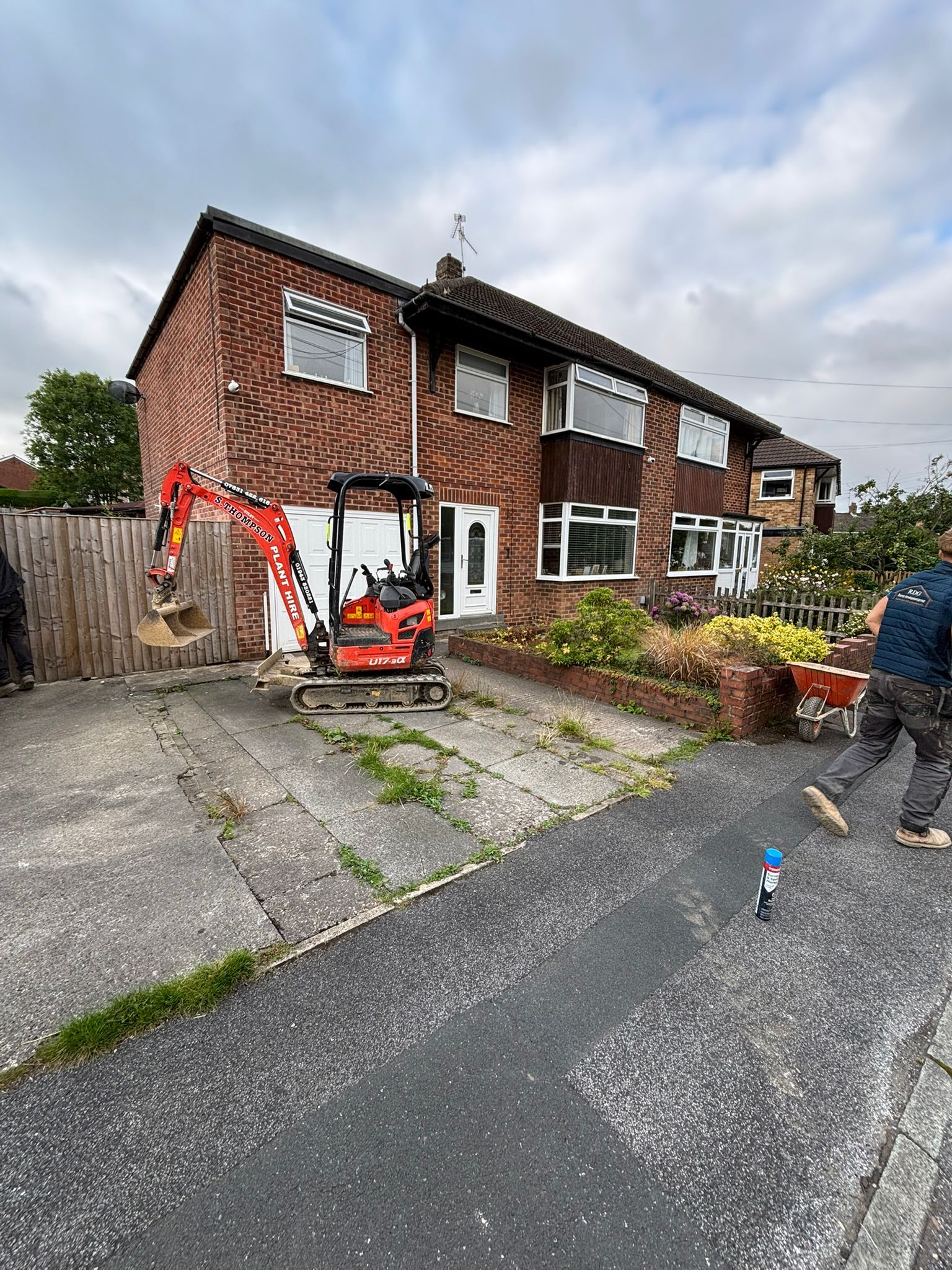 Brick semi-detached house with orange digger on the driveway and a person standing by the curb