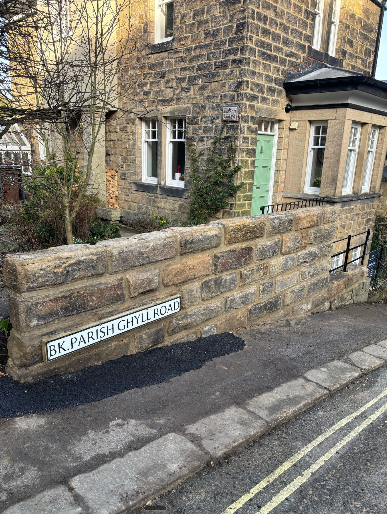 Stone wall with street sign reading “BAGPIPER” beside a stone house on a narrow road.