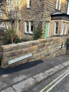 Stone wall with street sign reading “BAGPIPER” beside a stone house on a narrow road.