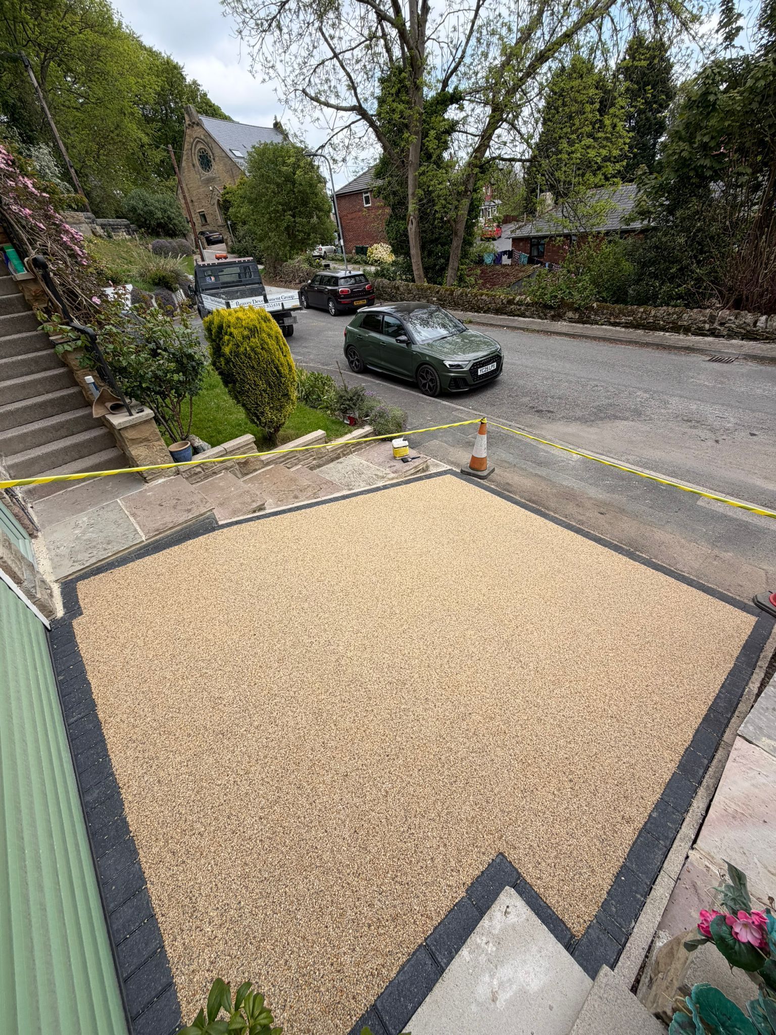 Gravel driveway and street outside a stone house, with parked cars and trees lining the road.