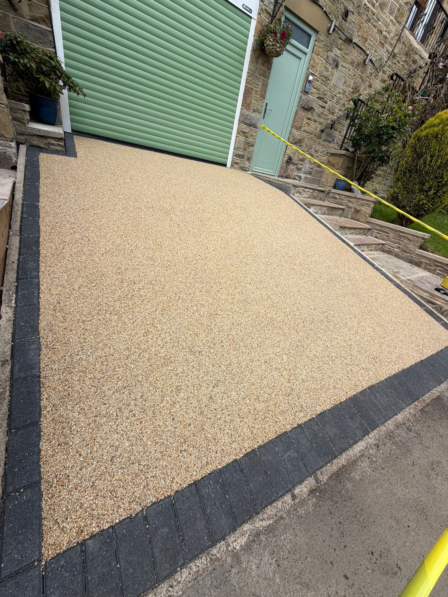 Tan gravel driveway beside a green garage door and stone wall, bordered by dark paving strips.