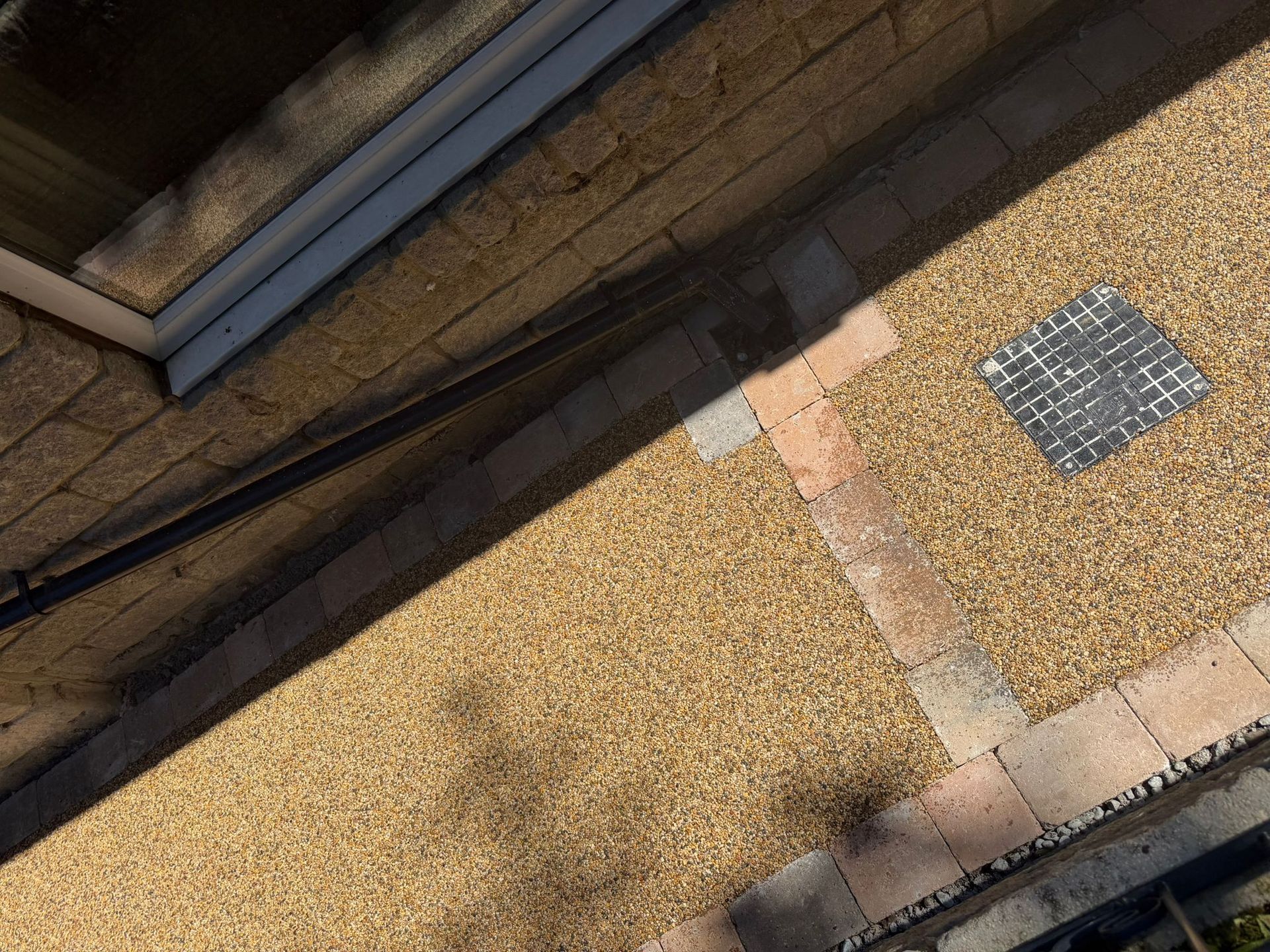 Slanted view of a gravel driveway with a brick border, metal drain grate, and building edge at top left