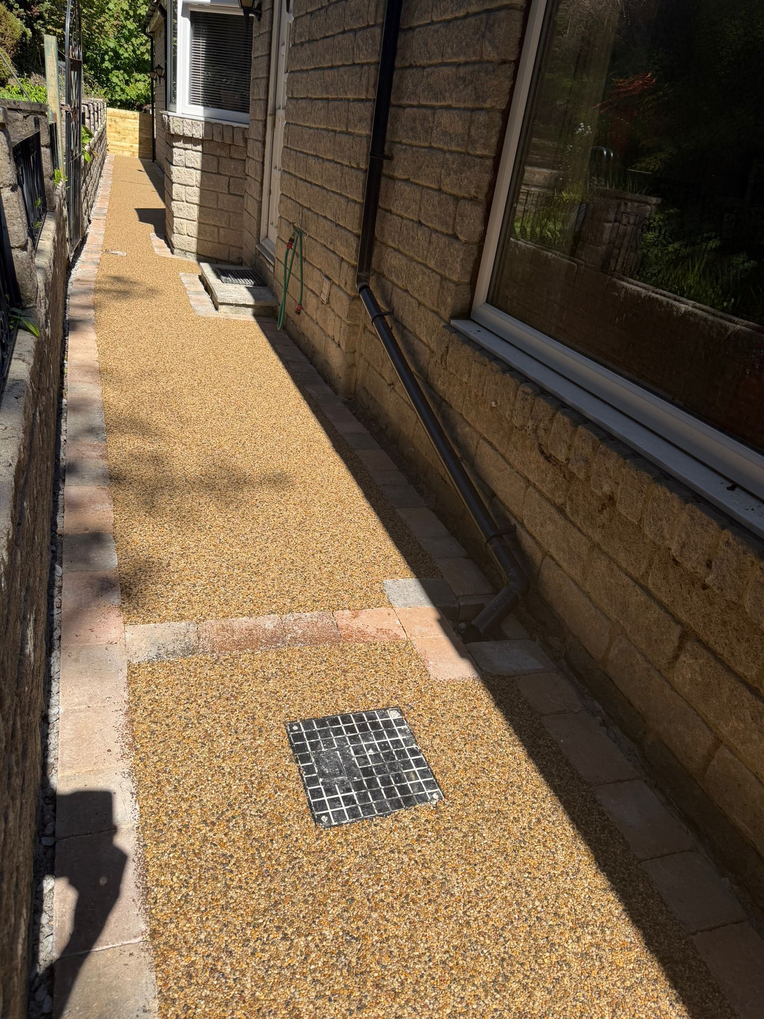 Narrow gravel walkway beside a brick house, with a small drain cover and fence in bright sunlight.