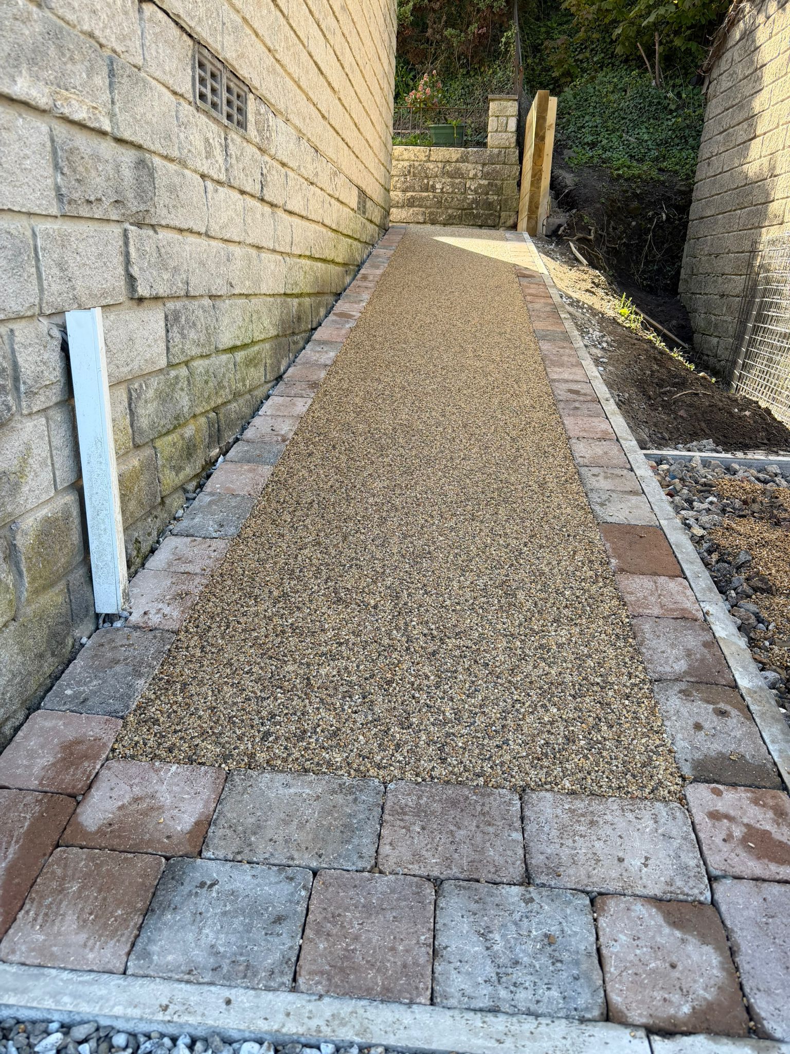 Gravel pathway between stone walls, bordered by pavers and a white post, leading uphill into sunlight