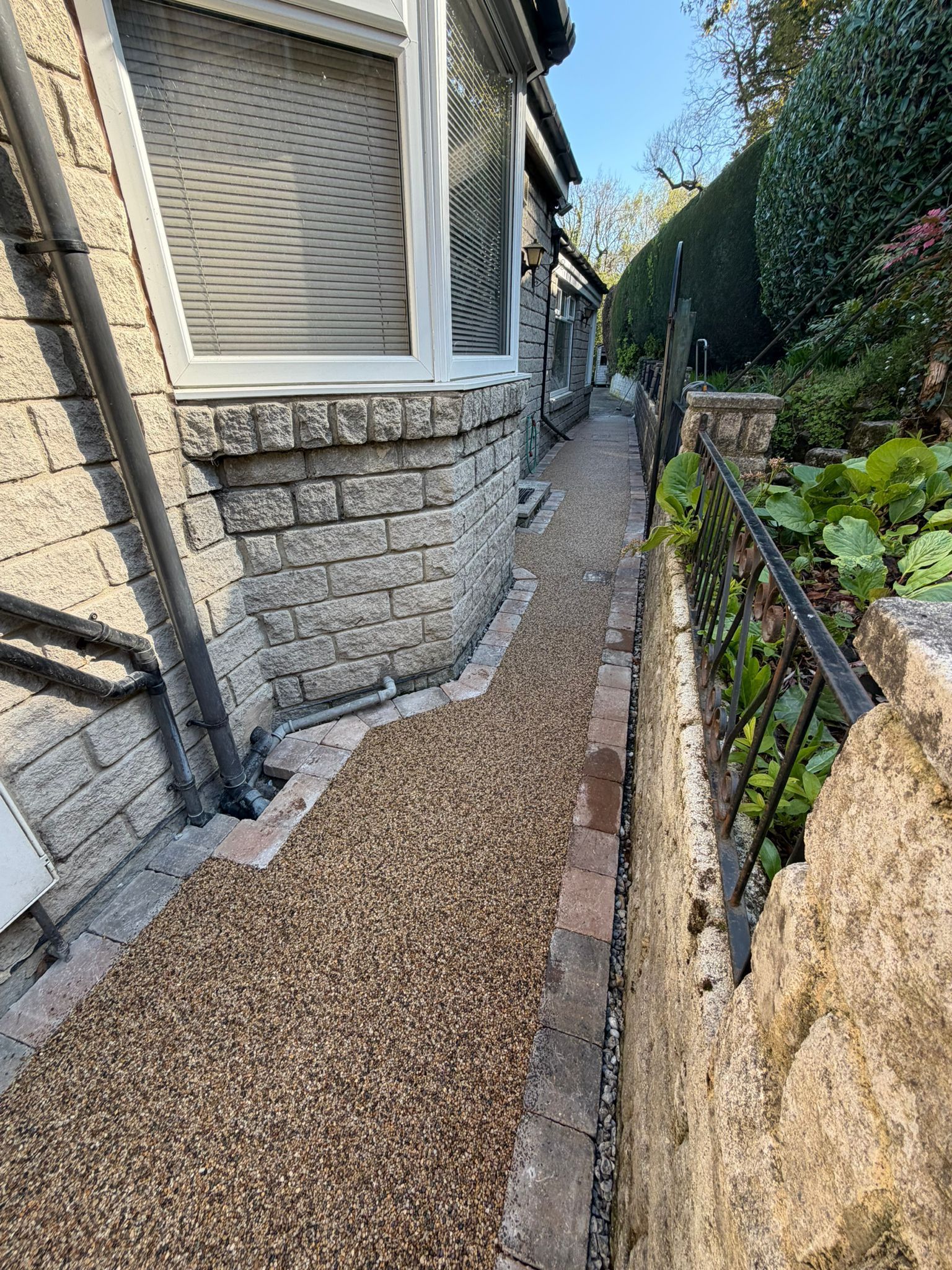 Narrow gravel path beside a stone house and garden wall, with hedges and steps ahead.